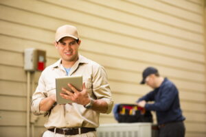 Latin descent repairman works on a home's air conditioner unit outdoors. One foreground is using a digital tablet. Other man in backgroud is checking unit using the tools in his tool bag.  They both wear uniforms.  Repairmen, electricians, inspectors, service workers.